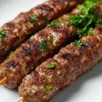 Close-up of a juicy grilled beef kofta kebab, garnished with parsley, placed on a plain white surface.
