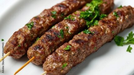 Close-up of a juicy grilled beef kofta kebab, garnished with parsley, placed on a plain white surface.